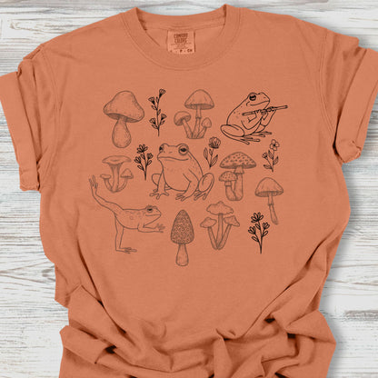 Brown t-shirt with frog and mushroom design on a wooden surface