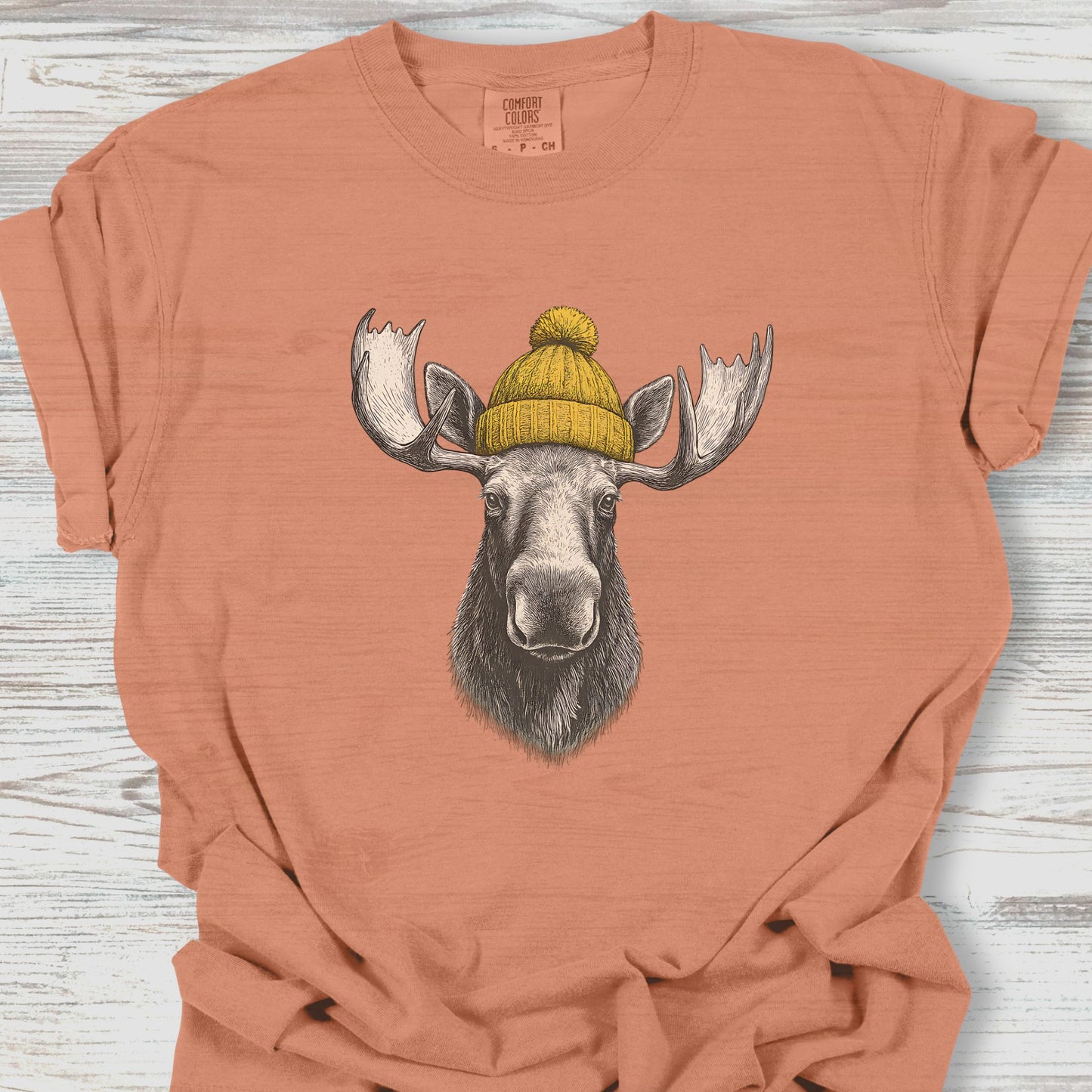 Coral t-shirt with a moose wearing a yellow beanie on a wooden surface