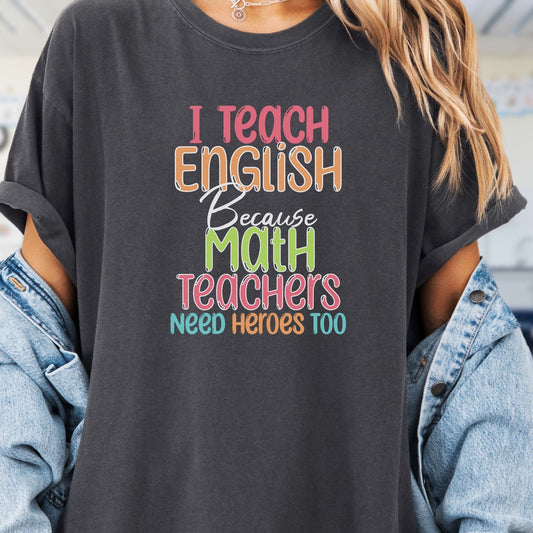 Model wears a dark grey tshirt with fun pastel rainbow lettering in a chunky font that reads, I Teach English because math teachers need heroes too.  Would make a great gift for an English Teacher.