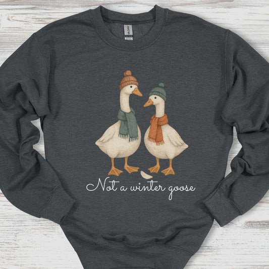 Dark gray sweatshirt with two geese wearing hats and scarves, and text 'Not a winter goose' on a wooden surface.