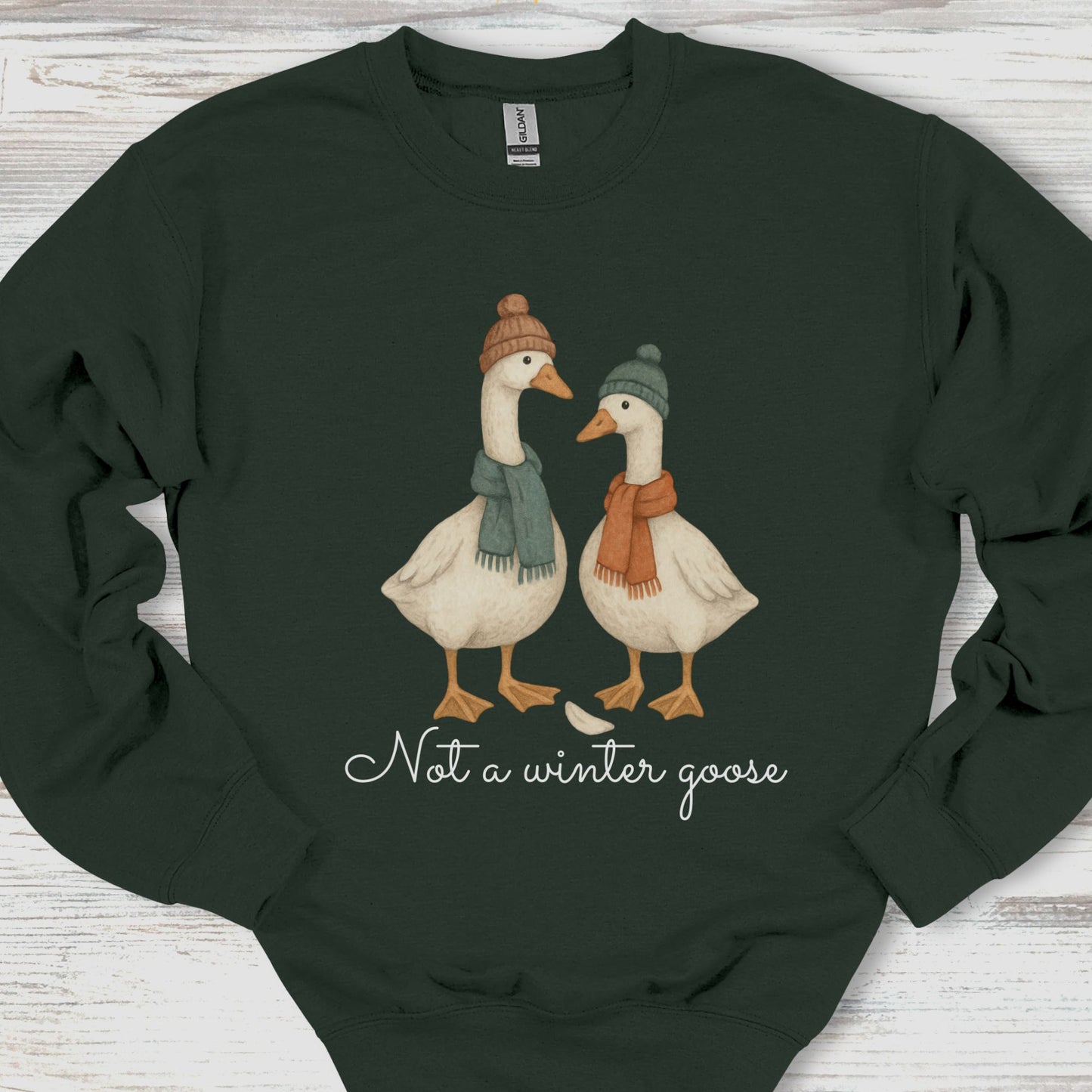 Dark green sweatshirt with two illustrated geese wearing hats and scarves, and text 'Not a winter goose' on a wooden surface.