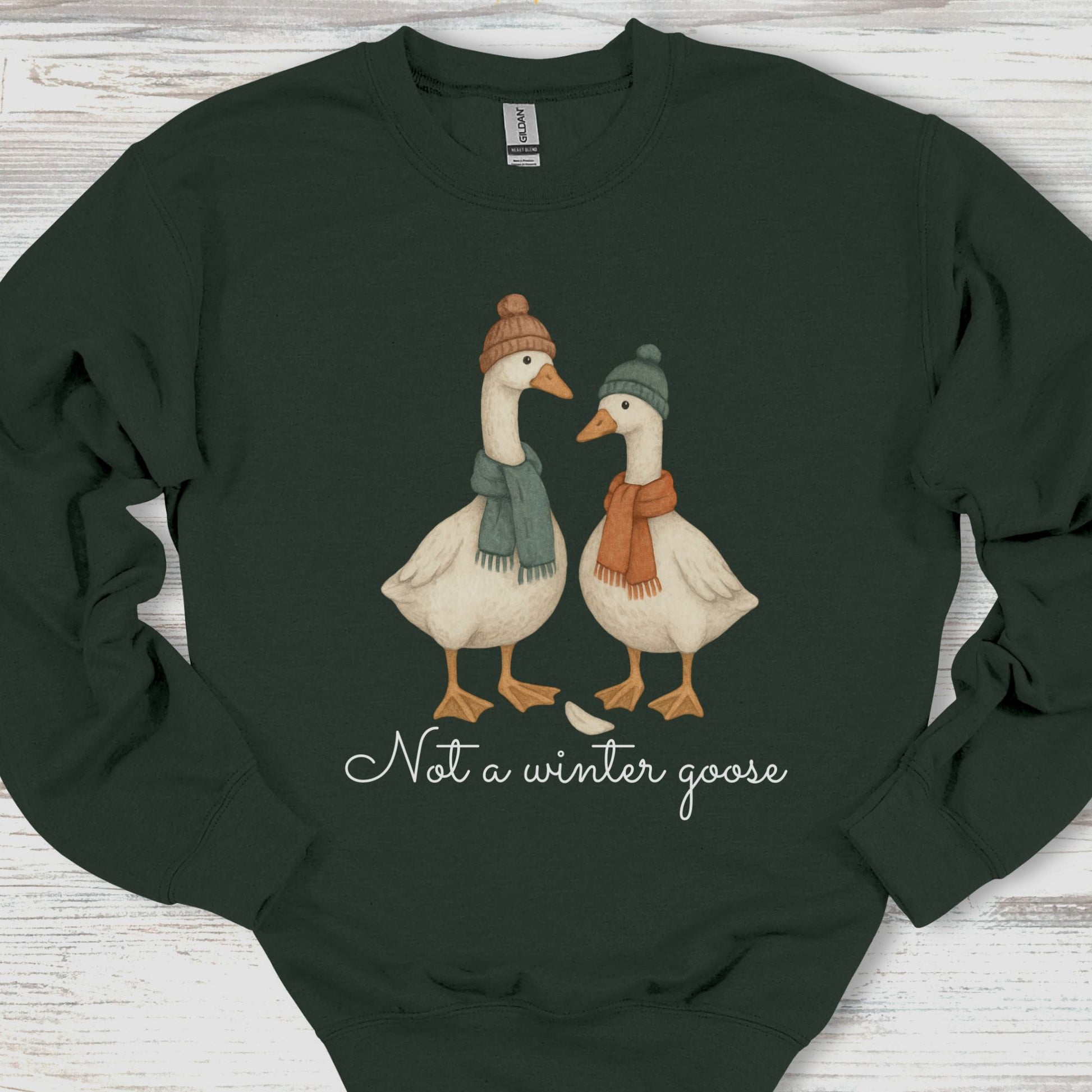 Dark green sweatshirt with two illustrated geese wearing hats and scarves, and text 'Not a winter goose' on a wooden surface.