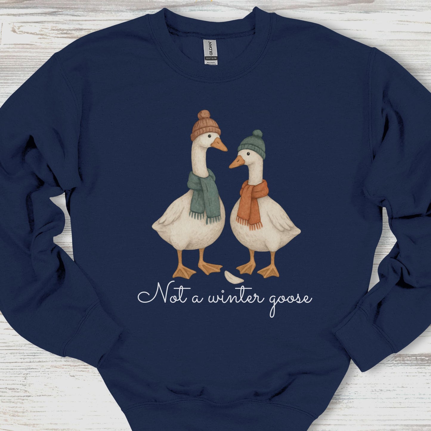 Navy blue sweatshirt with two illustrated geese wearing hats and scarves, and text 'Not a winter goose' on a wooden surface.