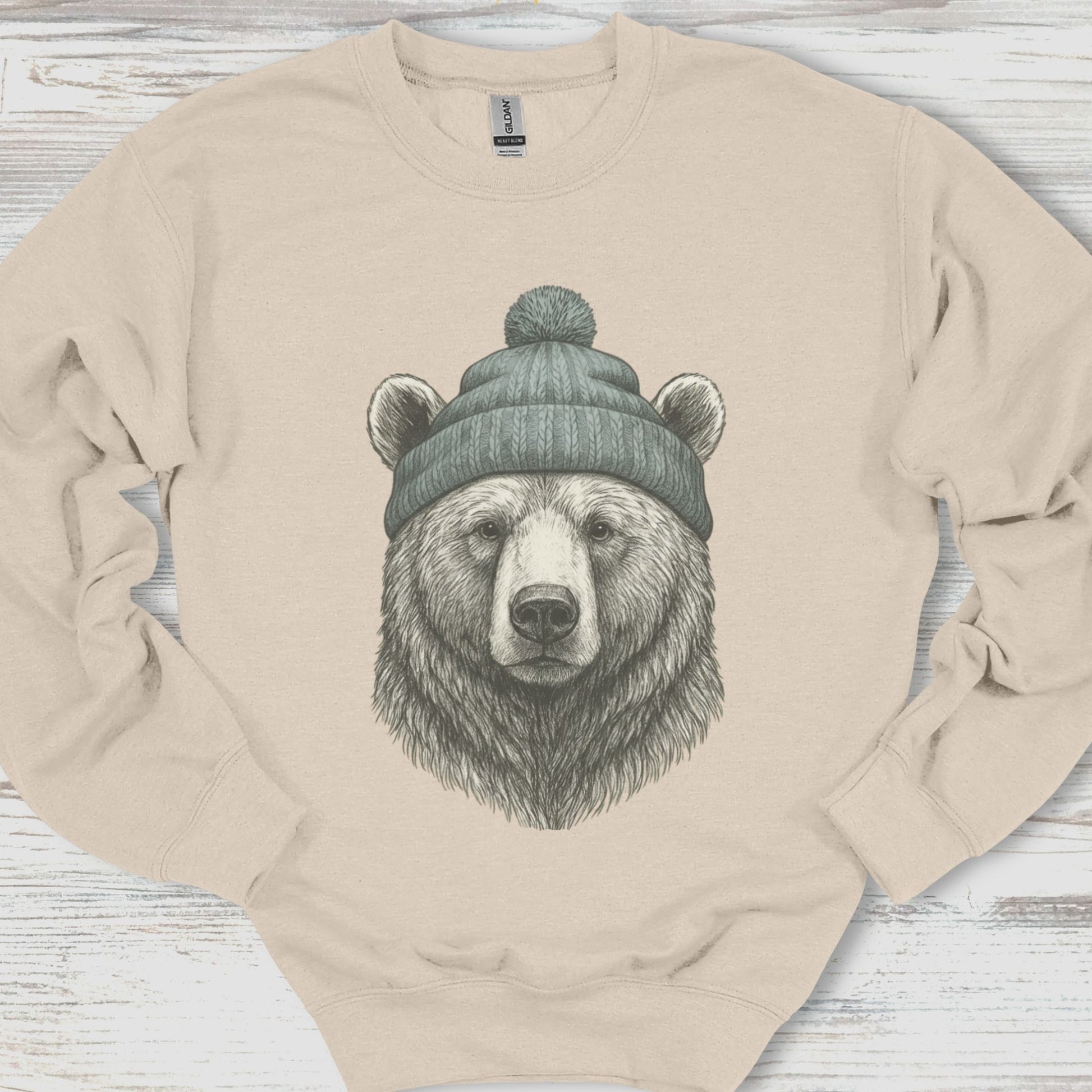 Beige sweatshirt with a bear wearing a beanie design on a wooden surface