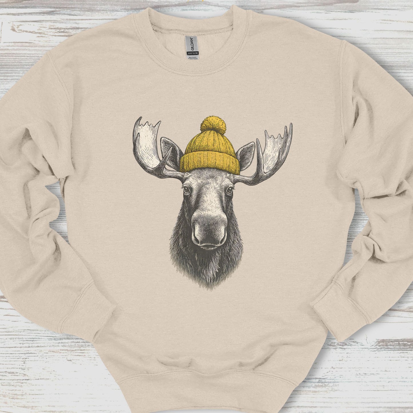 Beige sweatshirt with a moose wearing a yellow beanie on a wooden surface