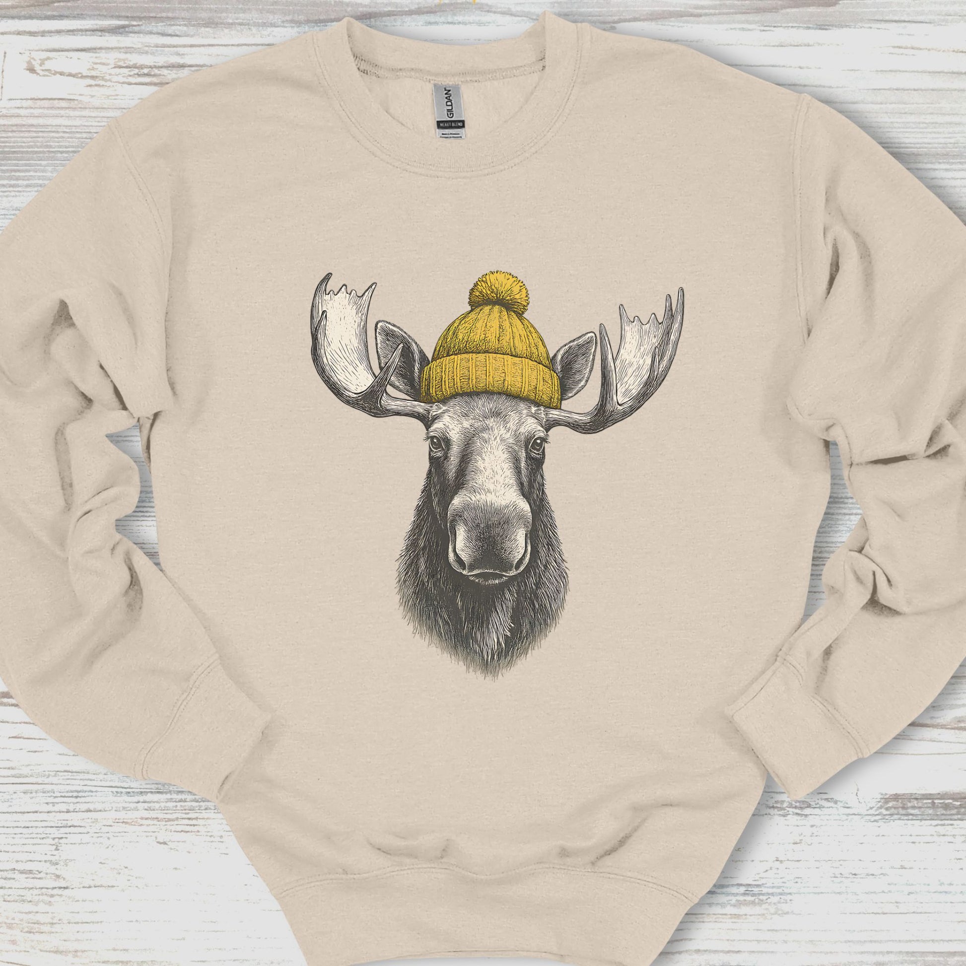 Beige sweatshirt with a moose wearing a yellow beanie on a wooden surface