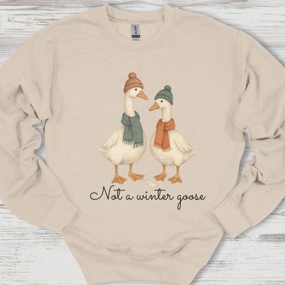 Beige sweatshirt with cartoon geese wearing hats and scarves and text 'Not a winter goose' on a wooden surface.