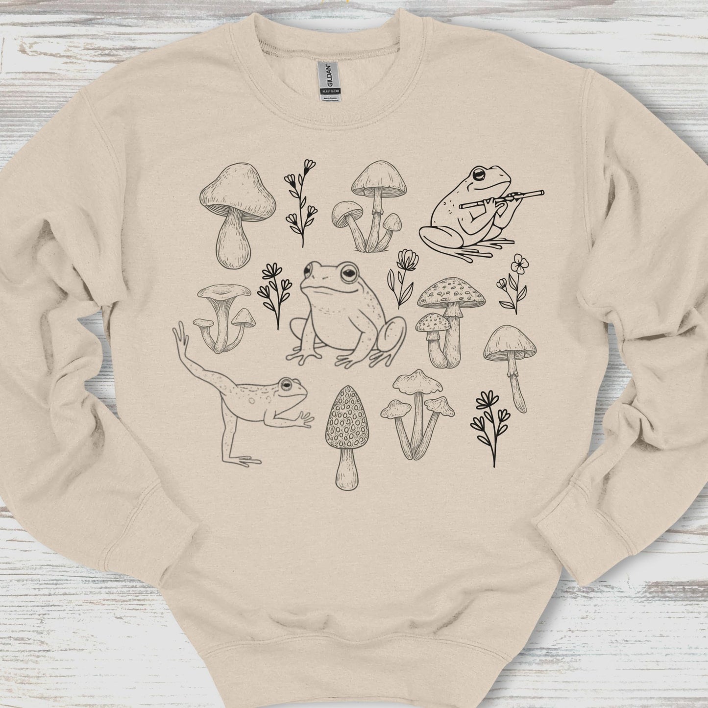 Beige sweatshirt with black line art of frogs, mushrooms, and plants on a wooden surface