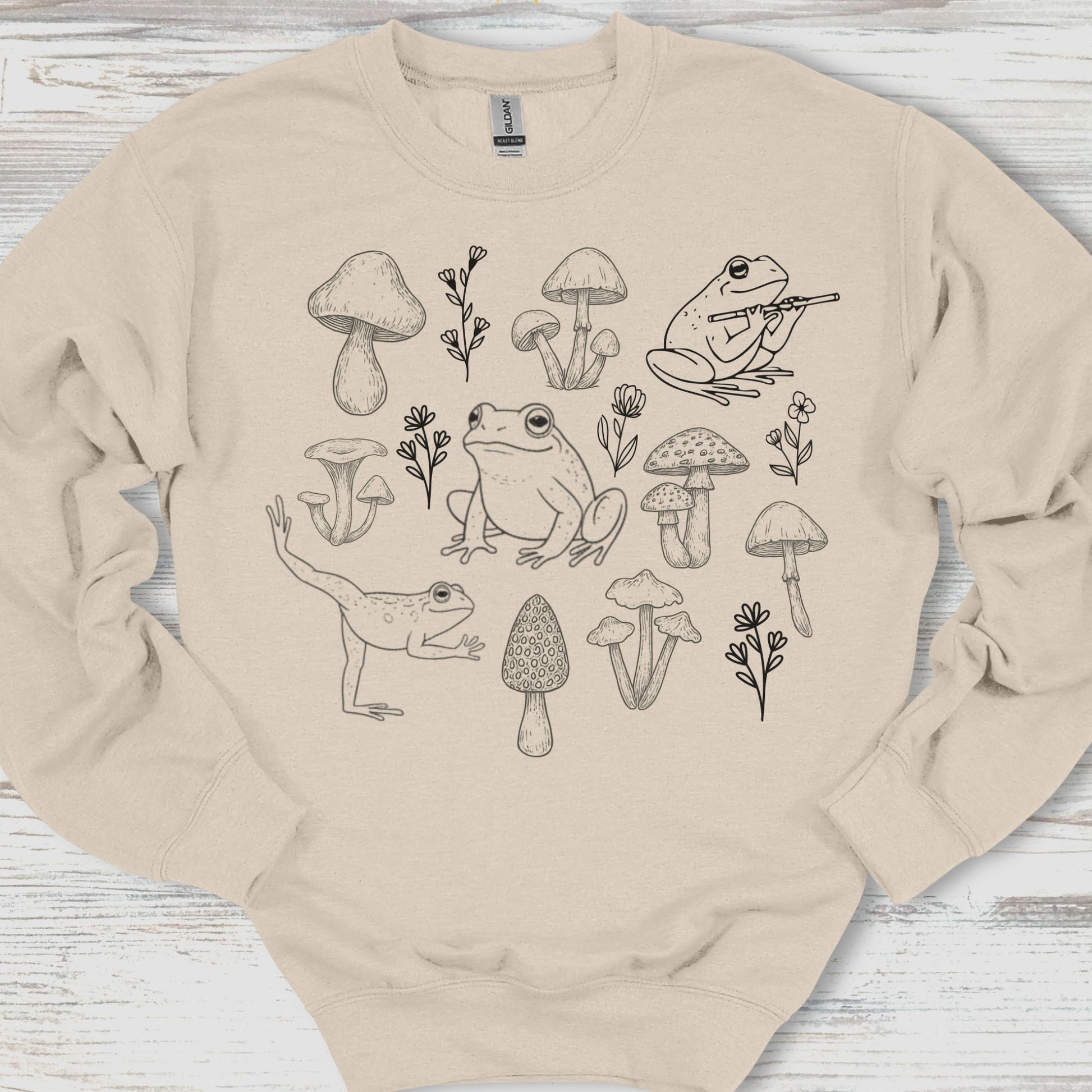 Beige sweatshirt with black line art of frogs, mushrooms, and plants on a wooden surface
