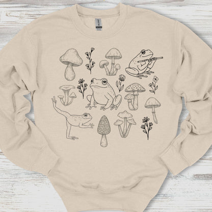 Beige sweatshirt with black line art of frogs, mushrooms, and plants on a wooden surface
