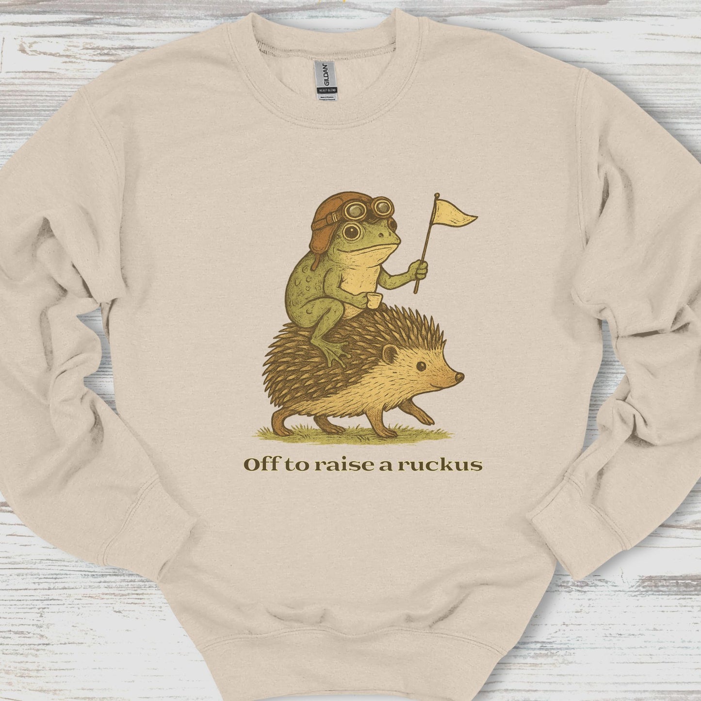 Beige sweatshirt with a graphic of a frog riding a hedgehog and text 'Off to raise a ruckus' on a wooden surface.