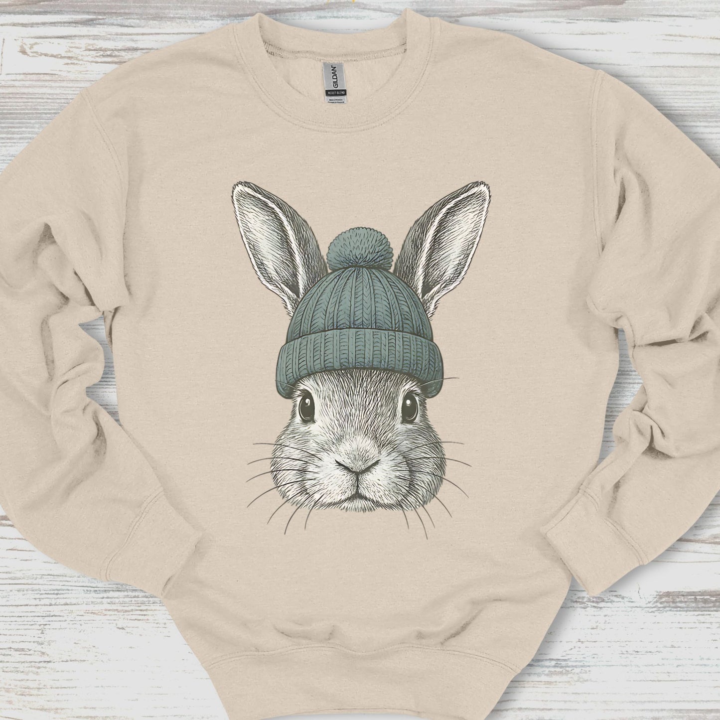 Beige sweatshirt with a graphic of a rabbit wearing a green beanie on a wooden surface