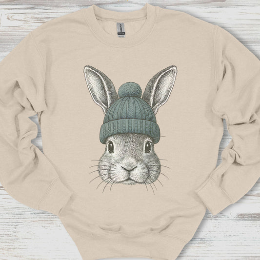 Beige sweatshirt with a graphic of a rabbit wearing a green beanie on a wooden surface