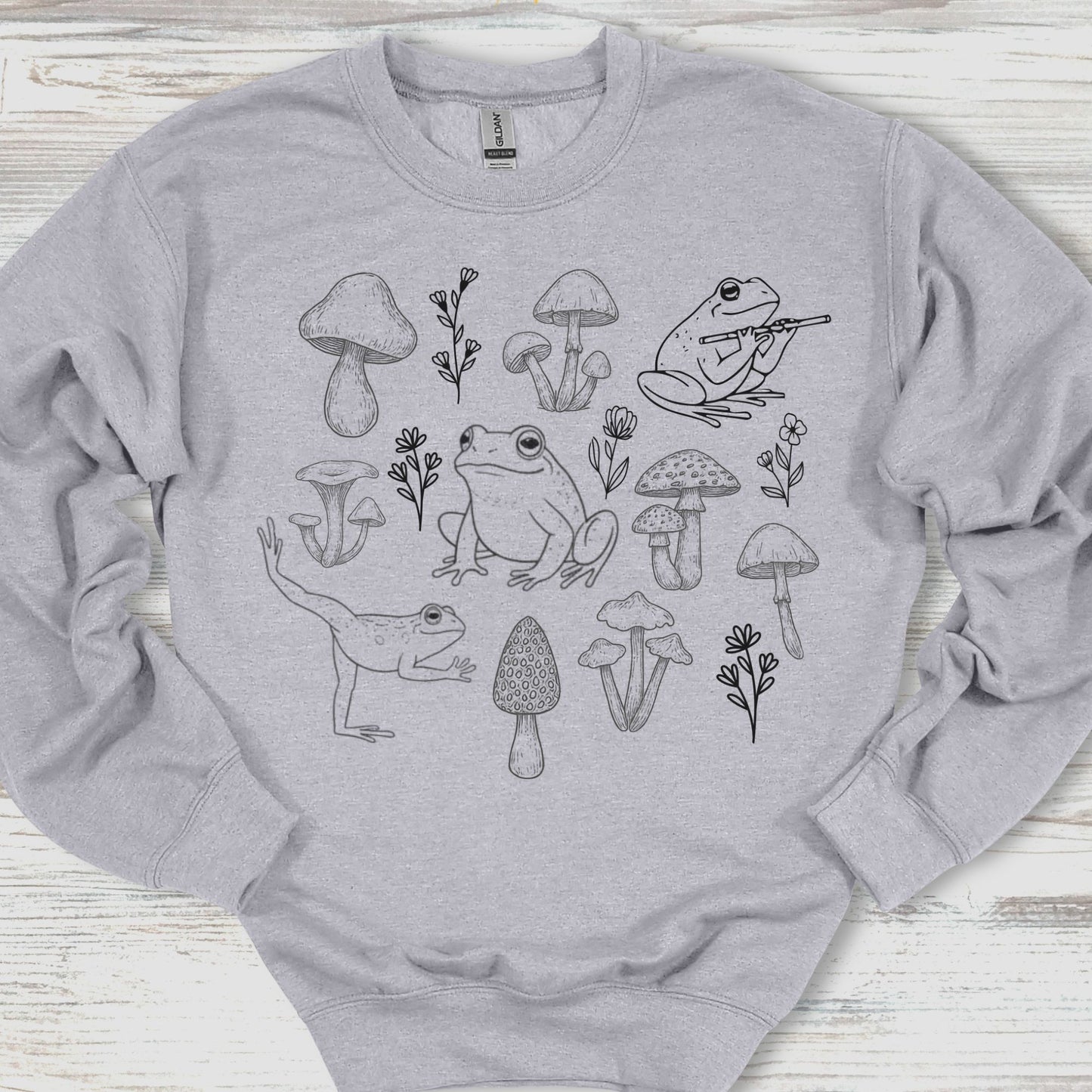 Gray sweatshirt with black line art of frogs and mushrooms on a wooden surface