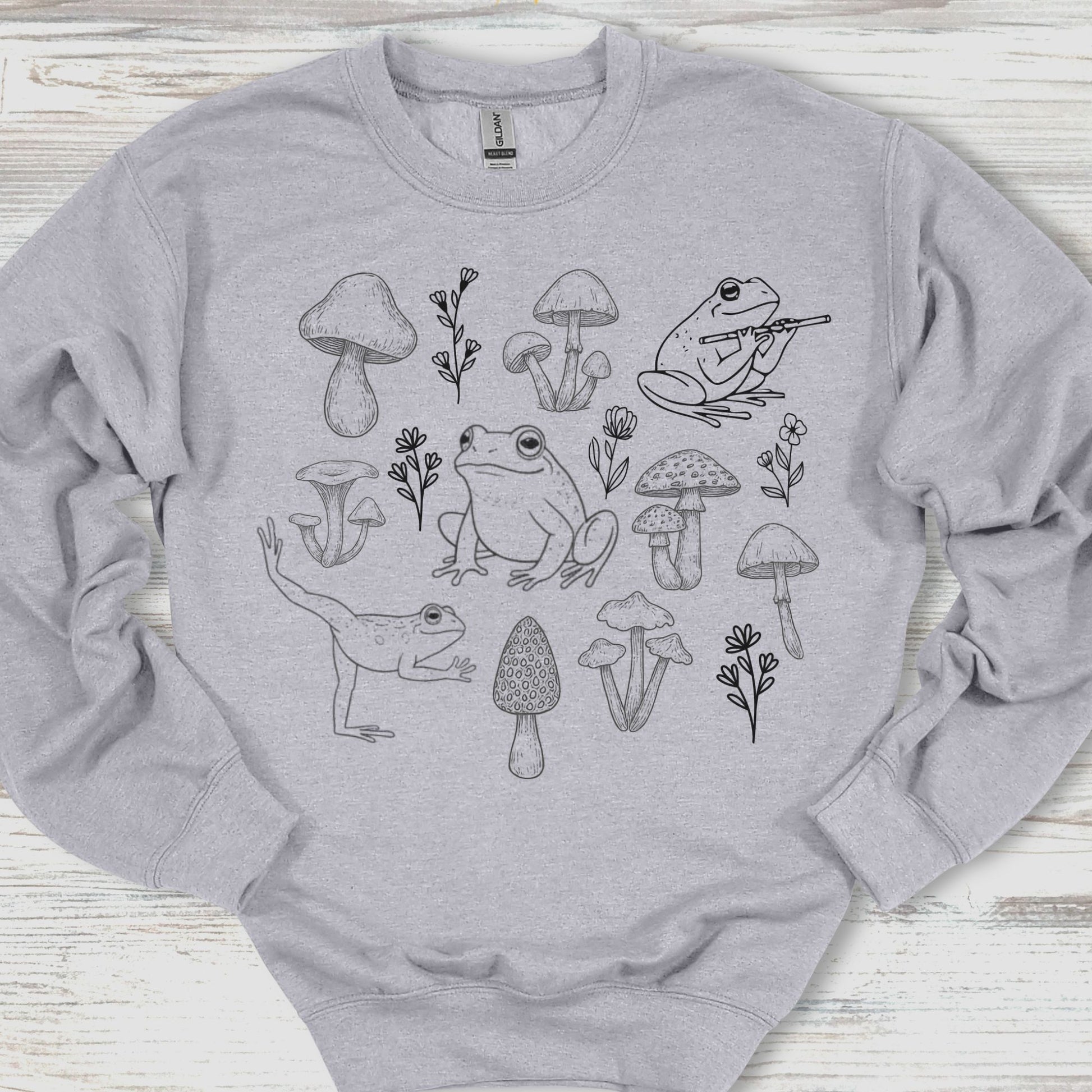 Gray sweatshirt with black line art of frogs and mushrooms on a wooden surface