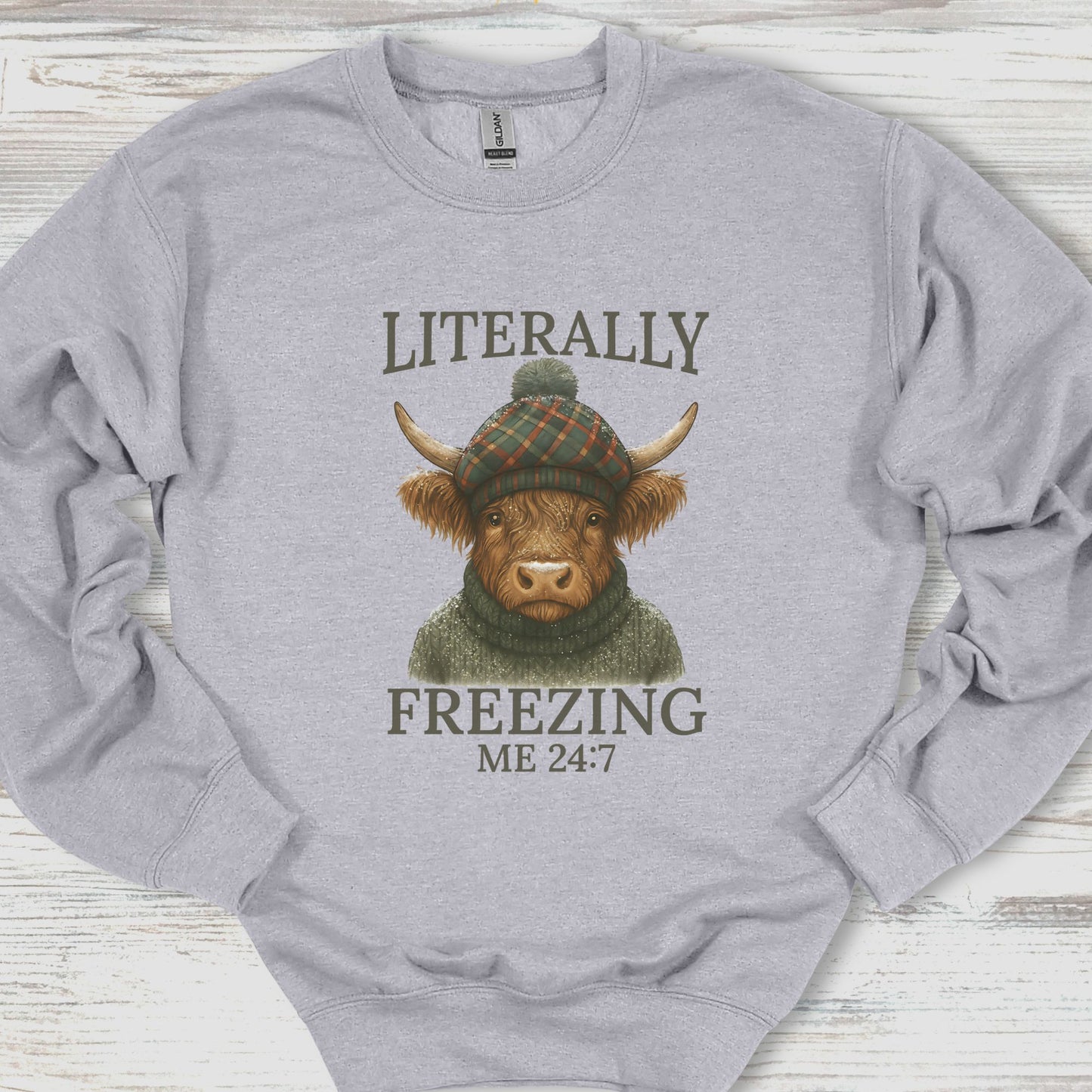 Gray sweatshirt with a graphic of a cow wearing a hat and sweater, text 'Literally Freezing Me 24:7', on a wooden surface.
