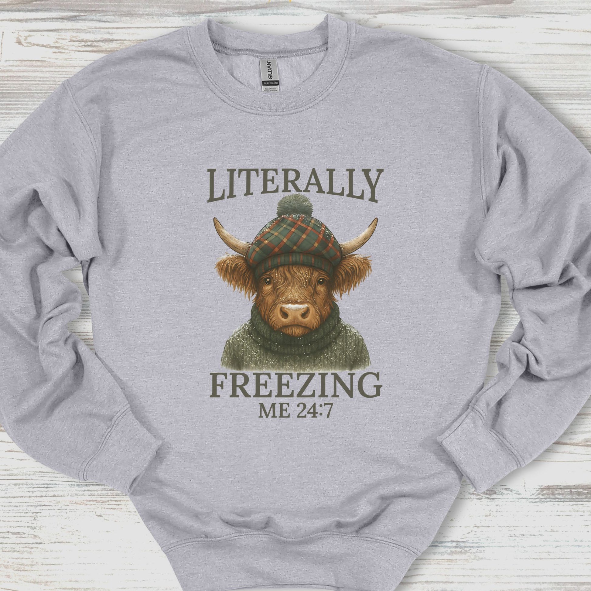 Gray sweatshirt with a graphic of a cow wearing a hat and sweater, text 'Literally Freezing Me 24:7', on a wooden surface.