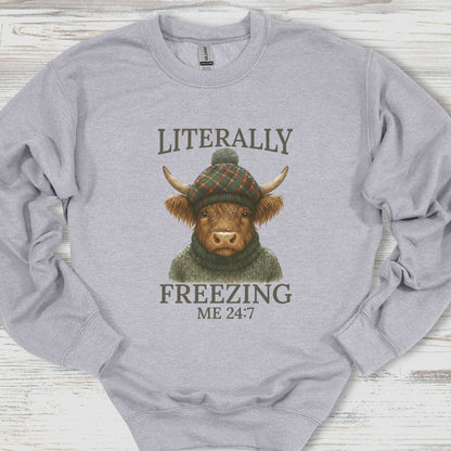 Gray sweatshirt with a graphic of a cow wearing a hat and sweater, text 'Literally Freezing Me 24:7', on a wooden surface.