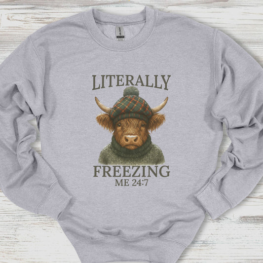 Gray sweatshirt with a graphic of a cow wearing a hat and sweater, text 'Literally Freezing Me 24:7', on a wooden surface.