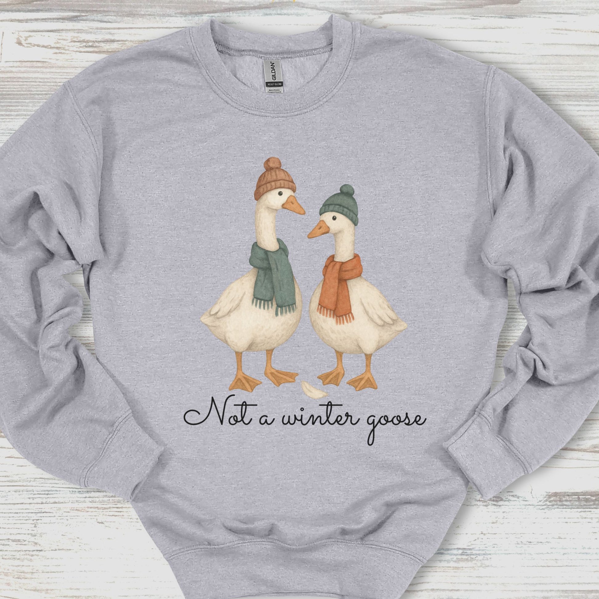 Gray sweatshirt with two geese wearing hats and scarves, and text 'Not a winter goose' on a wooden surface.