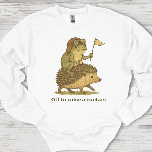 White sweatshirt with a graphic of a frog riding a hedgehog and text 'Off to raise a ruckus' on a wooden surface.