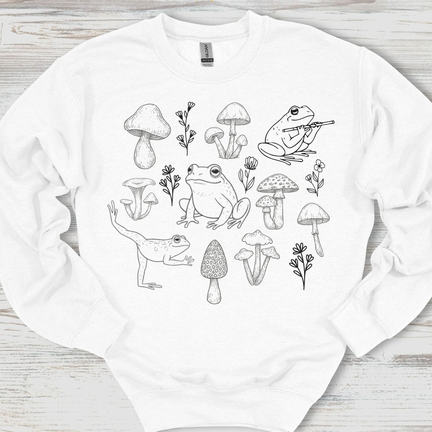 White sweatshirt with black line art design of frogs and mushrooms on a wooden surface