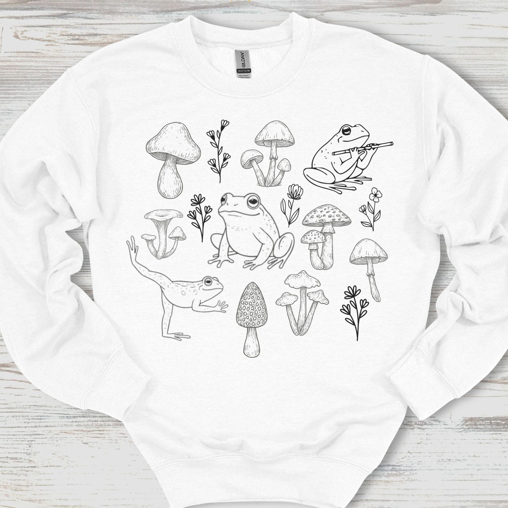 White sweatshirt with black line art design of frogs and mushrooms on a wooden surface