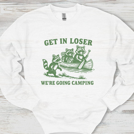 White sweatshirt with green graphic and text 'Get in Loser, We're Going Camping' on a wooden surface