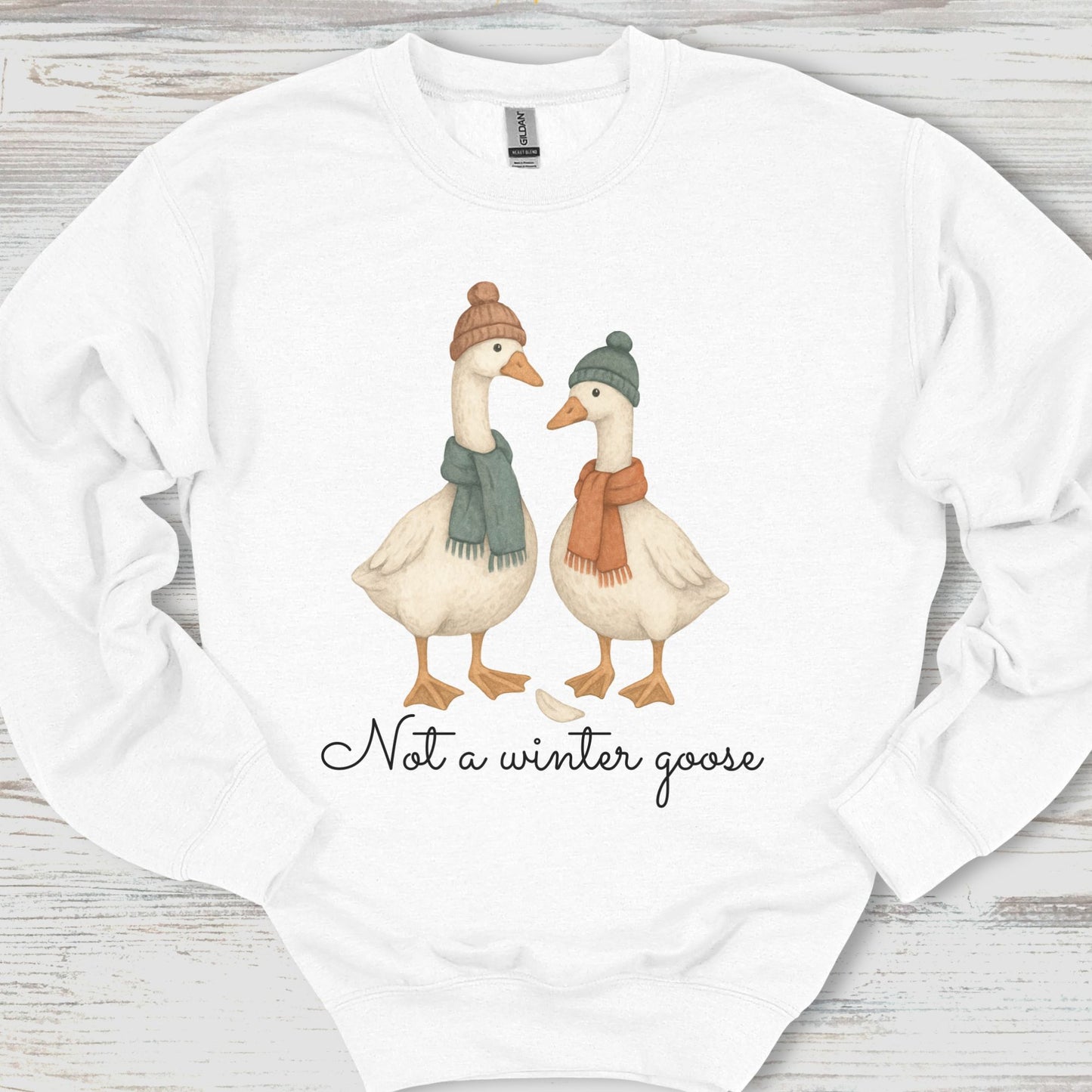 White sweatshirt with two cartoon geese wearing hats and scarves, and text 'Not a winter goose' on a wooden surface.