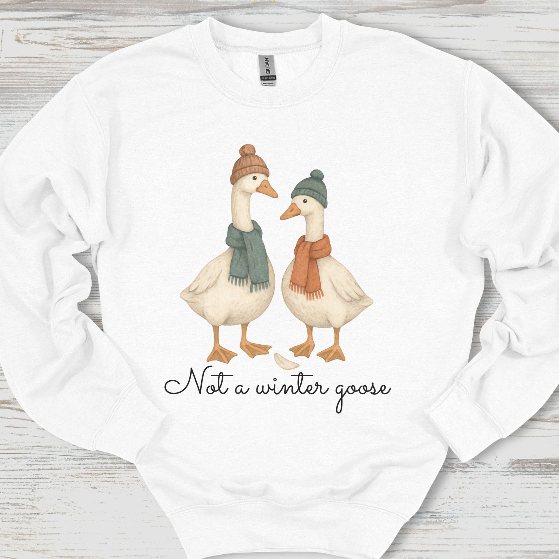 White sweatshirt with two cartoon geese wearing hats and scarves, and text 'Not a winter goose' on a wooden surface.