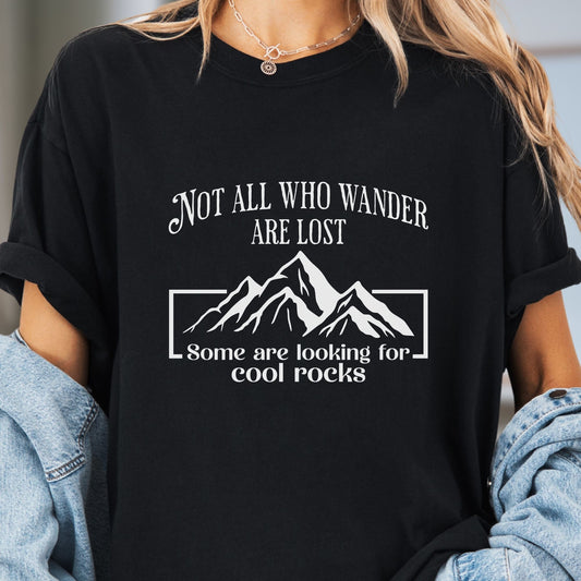 Model wears a black tshirt with white graphic of a mountain range and white lettering that reads Not all who wander are lost, some are looking for cool rocks.  Makes a great gift for a science teacher or geology lover.